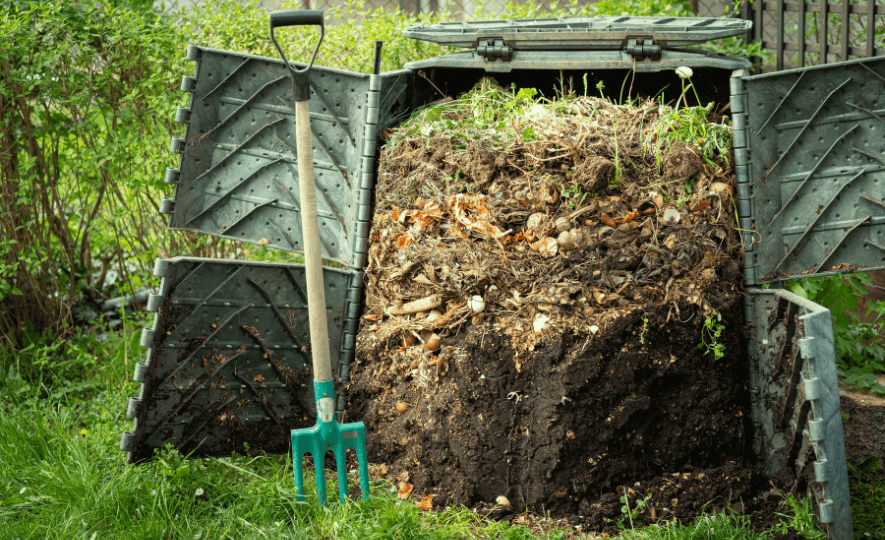 Compost bin filled with organic waste and decaying materials, with a garden fork leaning against it, in a backyard garden setting. Composting Organic Waste