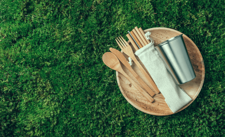 Reusable Serveware and Tableware set including bamboo utensils, reusable straws in a cloth pouch, and a stainless steel cup, placed on a wooden plate over a green grassy background.