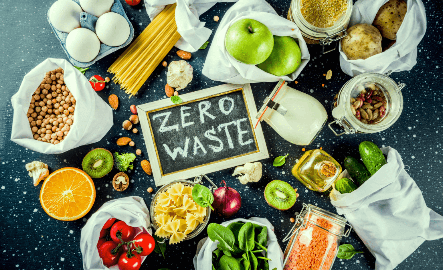 Zero-Waste Menu featuring fresh fruits, vegetables, pasta, lentils, chickpeas, and nuts in reusable jars and cloth bags surrounding a chalkboard sign reading 'Zero Waste'.